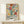 May include: Colorful abstract floral artwork in wooden frame leaning against white wall on light wood surface. Vibrant poster features botanical shapes in red, teal, yellow, purple, and orange with "Celebrating Colors of Life" text below. Small white gift box with twine sits in front. Matisse-inspired cut-out style with flowing organic forms and bold hues creates cheerful wall art display.