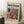 May include: Framed modernist art poster with bold abstract floral design in vibrant blues, oranges, reds, and yellows, displayed against white wall corner beside wooden side table with white coffee cup, sitting on textured cream rug next to beige throw blanket.