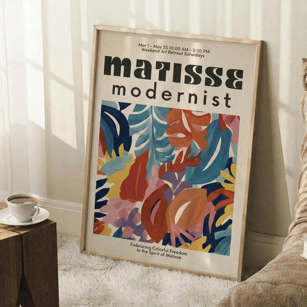 May include: Framed modernist art poster with bold abstract floral design in vibrant blues, oranges, reds, and yellows, displayed against white wall corner beside wooden side table with white coffee cup, sitting on textured cream rug next to beige throw blanket.
