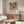 May include: Colorful abstract botanical poster with tropical leaves, fruits and plants in vibrant blues, oranges, greens and pinks, displayed in thin wooden frame above mid-century walnut credenza. Modern living room setting features white curved chair, glass oval coffee table, and natural lighting creating warm shadows on neutral walls.