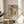 May include: Framed colorful abstract floral art print displayed on round wooden side table next to beige upholstered chair. Artwork features vibrant botanical shapes in coral, teal, yellow, and pink with "Celebrating Colors of Life" text below. Natural lighting creates soft shadows across white wall background with plant leaves visible in corner.