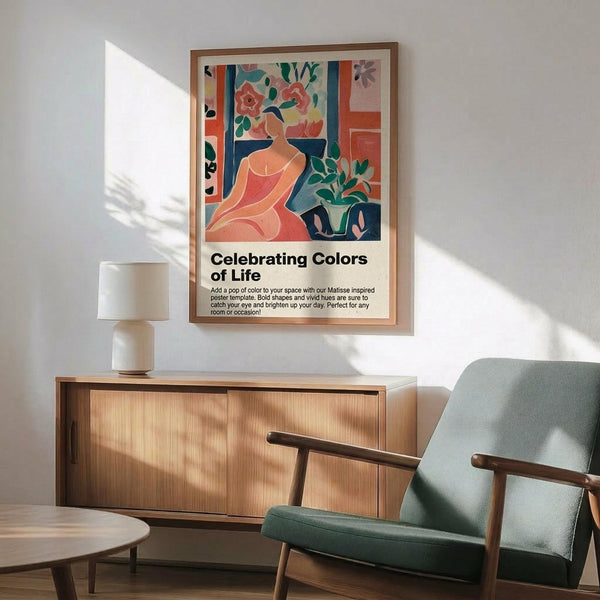 May include: A framed poster featuring a colorful Matisse-style illustration of a woman in coral dress surrounded by vibrant flowers and plants in blues, greens, and warm tones. The poster has "Celebrating Colors of Life" text below the artwork. The frame appears light wood-toned, hanging above a mid-century modern wooden sideboard with a white ceramic lamp. A sage green upholstered chair with wooden arms sits nearby on a wooden floor with natural sunlight creating shadows on the white wall.