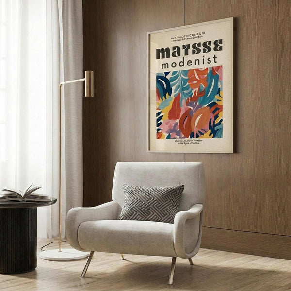 May include: Cream armchair with geometric patterned pillow positioned next to wood paneled wall. Framed colorful abstract art poster featuring flowing organic shapes in blues, oranges, and reds with "matisse modernist" text. Brass floor lamp and dark side table with open book complete the modern living room setting with natural lighting.