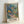 May include: Framed art print featuring a vibrant jungle scene with lush tropical foliage in blues, greens, oranges and purples. A small figure walks along a winding path through dense vegetation with palm fronds and colorful flowering plants. The artwork has a painterly, impressionistic style with layered colors and organic shapes. Light natural wood frame displays against white wall, leaning on surface with soft shadows.