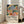 May include: Colorful abstract botanical poster in light wood frame leaning against white wall next to beige couch. Artwork features vibrant blues, greens, oranges, and pinks in painterly style with tropical leaves, flowers, and organic shapes. Text below reads "Celebrating Colors of Life" with description. Bright, airy room with large windows and natural lighting.