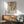May include: Colorful abstract floral poster with Matisse-inspired cut-out style flowers and leaves in vibrant reds, greens, yellows, and purples, displayed in thin wooden frame above mid-century modern wooden sideboard with white ceramic lamp, next to sage green upholstered armchair with wooden arms in bright sunlit room with white walls and natural shadows
