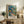 May include: Framed art print featuring vibrant tropical garden scene with colorful flowers, palm fronds, and foliage in blues, oranges, and greens, displayed on rustic wooden sideboard alongside fern plant, ceramic vase, and books in cozy living room setting with cream armchair and neutral walls.