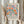May include: Two hands holding a framed botanical poster with colorful stylized flowers in orange, pink, blue and green against cream background. The frame has light wood finish and poster reads "Celebrating Colors of Life" with Lorem ipsum text below. Dried pampas grass visible on left side against neutral beige curtained background.