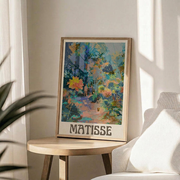 May include: Colorful tropical garden scene poster with "MATISSE" text below the artwork, displayed in a thin wooden frame on a round wooden side table next to a beige upholstered chair. The vibrant print features lush jungle foliage in blues, greens, yellows, and coral tones with a winding path through dense vegetation. Natural sunlight streams across the neutral-toned room setting.