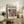 May include: Framed modernist art poster with abstract colorful shapes in blues, reds, oranges and yellows, displayed on rustic wooden sideboard alongside fern plant, ceramic vases, and books in cozy living room with cream armchair and neutral walls