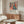 May include: Matisse-inspired colorful poster featuring woman in coral dress surrounded by flowers and plants, displayed in wooden frame above mid-century walnut sideboard. Modern living room with cream curved chair, glass oval coffee table, and neutral walls. Artwork shows bold geometric shapes in blues, corals, greens with "Celebrating Colors of Life" text below illustration.