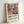 May include: Framed poster print leaning against white wall showing colorful Matisse-inspired artwork of woman in coral dress surrounded by flowers and plants in blues, greens, oranges and pinks. Light wood frame with white mat. Text below reads "Celebrating Colors of Life" with description about adding color to spaces.