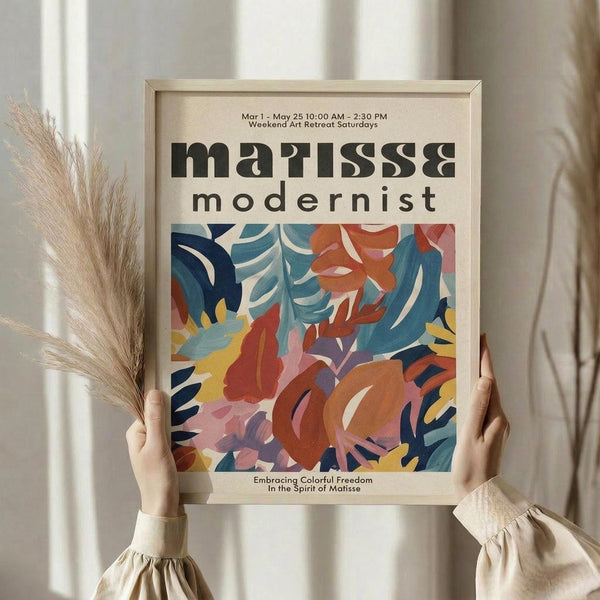 May include: Person in beige ruffled sleeves holding framed poster against white curtained window, with dried pampas grass visible to left. Poster features vibrant abstract cutout design in blues, oranges, pinks and yellows reminiscent of Matisse's paper collage style, with black text reading "matisse modernist" and event details above colorful botanical shapes in light wood frame.