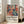 May include: Framed poster with Matisse-inspired artwork showing a seated woman in coral dress surrounded by colorful flowers and plants in blues, greens, and oranges. Text below reads "Celebrating Colors of Life" with descriptive paragraph. Light wood frame leaning against white wall next to beige sofa in bright, airy room with large windows and hardwood floors.