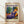 May include: Framed poster featuring a colorful Matisse-style painting of a reclining woman in purple patterned clothing with orange headwrap, surrounded by potted plants and decorative objects in vibrant blues, greens, and yellows. The cream-colored poster displays "MATISSE" in bold purple letters at top, with "06200 NICE, FRANCE" and exhibition details at bottom. Natural wood frame sits against white wall on light wooden floor with soft shadows.