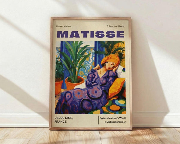 May include: Framed poster featuring a colorful Matisse-style painting of a reclining woman in purple patterned clothing with orange headwrap, surrounded by potted plants and decorative objects in vibrant blues, greens, and yellows. The cream-colored poster displays "MATISSE" in bold purple letters at top, with "06200 NICE, FRANCE" and exhibition details at bottom. Natural wood frame sits against white wall on light wooden floor with soft shadows.