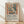 May include: Framed poster in light wood frame leaning against white textured wall on wooden floor. Features colorful Matisse-style interior scene with plant in vase, fruit bowl, and window view in warm earth tones of terracotta, sage green, and cream. Exhibition-style poster with black text below artwork describing artist's bold use of color and unique style. Soft natural lighting creates diagonal shadows across the scene.