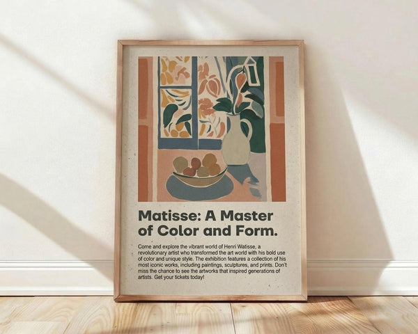 May include: Framed poster in light wood frame leaning against white textured wall on wooden floor. Features colorful Matisse-style interior scene with plant in vase, fruit bowl, and window view in warm earth tones of terracotta, sage green, and cream. Exhibition-style poster with black text below artwork describing artist's bold use of color and unique style. Soft natural lighting creates diagonal shadows across the scene.