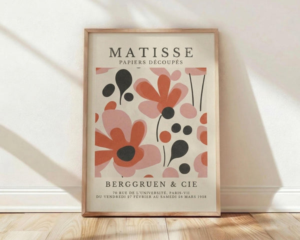May include: Framed vintage-style botanical art print featuring abstract coral pink and rust orange flowers with black stems and circles on cream background, displayed in light wood frame against white wall on wooden floor with natural lighting shadows