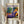 May include: Person in cream sweater holding framed Matisse exhibition poster featuring colorful Fauvist painting of reclining woman in purple patterned dress with orange turban, surrounded by potted plants. Poster shows blue text "MATISSE" at top with "Musees Matisse" and "Tribute to a Master" headers, location "06200 NICE, FRANCE" at bottom. Light wood frame, neutral background with pampas grass visible.