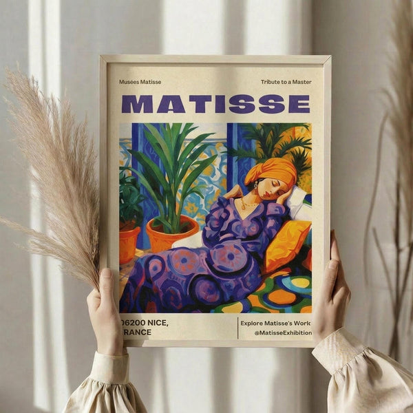 May include: Person in cream sweater holding framed Matisse exhibition poster featuring colorful Fauvist painting of reclining woman in purple patterned dress with orange turban, surrounded by potted plants. Poster shows blue text "MATISSE" at top with "Musees Matisse" and "Tribute to a Master" headers, location "06200 NICE, FRANCE" at bottom. Light wood frame, neutral background with pampas grass visible.