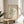May include: Framed art print featuring green organic coral-like shapes on cream background with "MATISSE PAPIERS DÉCOUPÉS" and gallery text, displayed on round wooden side table next to beige upholstered chair with plant leaves visible in corner, natural sunlight creating soft shadows on neutral walls.