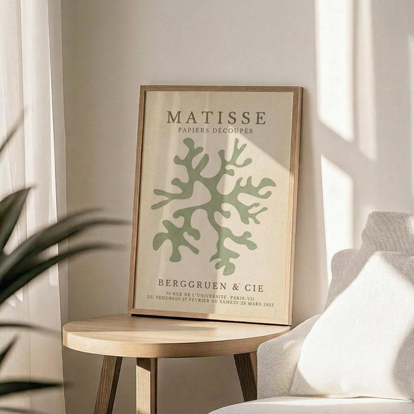 May include: Framed art print featuring green organic coral-like shapes on cream background with "MATISSE PAPIERS DÉCOUPÉS" and gallery text, displayed on round wooden side table next to beige upholstered chair with plant leaves visible in corner, natural sunlight creating soft shadows on neutral walls.