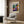 May include: colorful Matisse exhibition poster featuring woman in purple dress with plants and fruits, displayed in light wood frame on wood paneled wall above beige upholstered armchair with geometric patterned pillow, brass floor lamp and side table with open book nearby