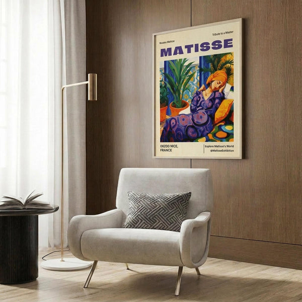 May include: colorful Matisse exhibition poster featuring woman in purple dress with plants and fruits, displayed in light wood frame on wood paneled wall above beige upholstered armchair with geometric patterned pillow, brass floor lamp and side table with open book nearby