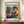 May include: Framed Matisse exhibition poster featuring a woman in purple patterned dress with orange head wrap, resting beside potted plants. Colorful artwork shows vibrant blues, oranges, and greens in Matisse's distinctive style. Wooden frame displayed on light wood sideboard next to white ceramic vase and stacked books in bright, minimalist room with white paneled walls.