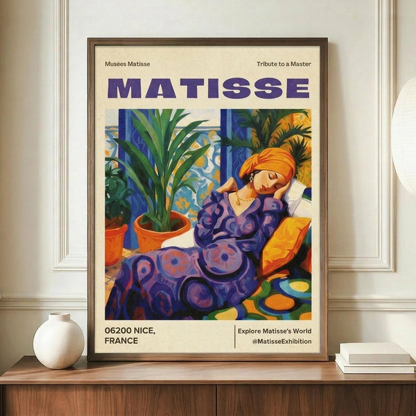 May include: Framed Matisse exhibition poster featuring a woman in purple patterned dress with orange head wrap, resting beside potted plants. Colorful artwork shows vibrant blues, oranges, and greens in Matisse's distinctive style. Wooden frame displayed on light wood sideboard next to white ceramic vase and stacked books in bright, minimalist room with white paneled walls.