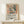 May include: Framed exhibition poster featuring Matisse-style still life with window, vase of flowers, and fruit bowl in warm earth tones and blues. Displayed on light wood surface alongside small wrapped gift with twine and greenery sprig against cream wall. Natural lighting creates soft shadows on the minimal modern interior setting.