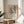 May include: Framed poster print featuring a Matisse-style interior scene with colorful flowers in a white vase, fruit bowl, and window view, displayed on a round wooden side table next to a beige upholstered chair with natural lighting casting window shadows across white walls and a plant in the corner