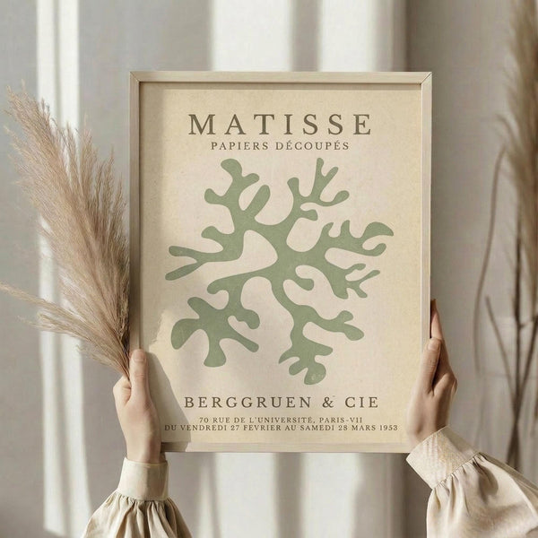 May include: Person in beige sweater holding framed poster featuring green coral-like abstract design on cream background, with "MATISSE PAPIERS DÉCOUPÉS" and French exhibition text. Dried pampas grass visible to left, soft natural lighting against white curtained window.