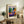 May include: Framed Matisse exhibition poster featuring a reclining woman in purple patterned clothing with vibrant orange hair, surrounded by tropical plants in terracotta pots. The artwork displays bold blues, purples, oranges and greens in Matisse's distinctive style. Shown in light wood frame on rustic wooden sideboard alongside fern plant, ceramic vase, and books in warm-toned living room setting.