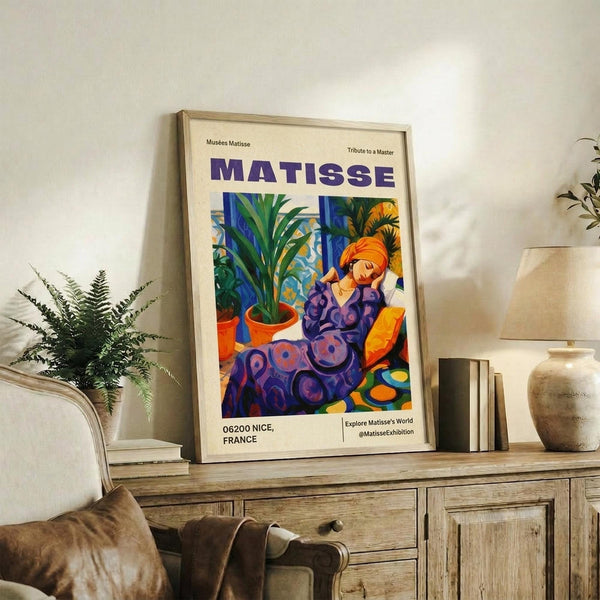 May include: Framed Matisse exhibition poster featuring a reclining woman in purple patterned clothing with vibrant orange hair, surrounded by tropical plants in terracotta pots. The artwork displays bold blues, purples, oranges and greens in Matisse's distinctive style. Shown in light wood frame on rustic wooden sideboard alongside fern plant, ceramic vase, and books in warm-toned living room setting.
