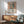 May include: Framed Matisse-style poster hanging above wooden sideboard in modern living room. Artwork features colorful still life with window view, vase with flowers, and fruit bowl in muted earth tones. Mid-century green armchair and white table lamp complete the scene with natural sunlight casting shadows on white wall.