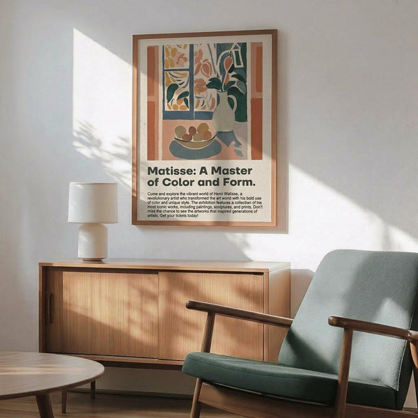 May include: Framed Matisse-style poster hanging above wooden sideboard in modern living room. Artwork features colorful still life with window view, vase with flowers, and fruit bowl in muted earth tones. Mid-century green armchair and white table lamp complete the scene with natural sunlight casting shadows on white wall.