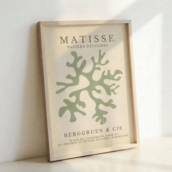 May include: Framed art print leaning against white wall showing Matisse-inspired paper cutout design with green coral-like organic shapes on cream background. Text reads "MATISSE PAPIERS DÉCOUPÉS" at top and "BERGGRUEN & CIE" with French exhibition details at bottom. Light wooden frame, soft natural lighting creates gentle shadows.