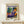 May include: Framed art poster featuring a woman in purple patterned dress with orange turban, reclining among tropical plants in terracotta pots. Bold colors include blues, greens, yellows and oranges in Matisse's distinctive style. Wooden frame sits on light wood surface next to small wrapped gift with twine and greenery sprig against cream wall.