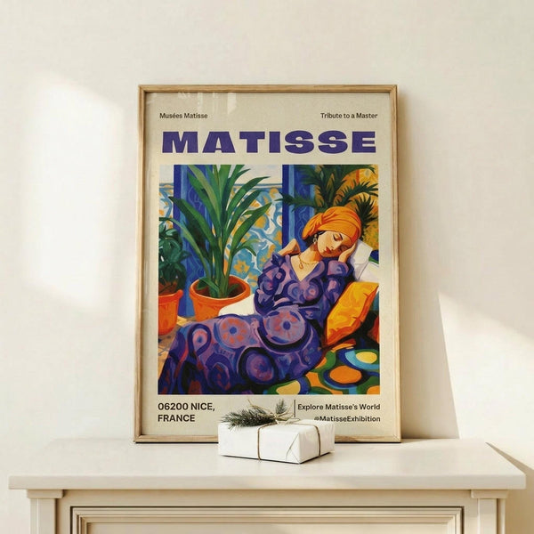 May include: Framed art poster featuring a woman in purple patterned dress with orange turban, reclining among tropical plants in terracotta pots. Bold colors include blues, greens, yellows and oranges in Matisse's distinctive style. Wooden frame sits on light wood surface next to small wrapped gift with twine and greenery sprig against cream wall.