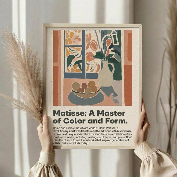 May include: Person in beige ruffled sleeves holding framed poster featuring Matisse-style still life artwork with vase of flowers, fruit bowl, and window view in warm earth tones of terracotta, sage green, and cream. Dried pampas grass visible beside figure against soft neutral curtained background. Exhibition-style poster with descriptive text below colorful geometric composition.