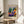 May include: Framed poster of Matisse's colorful artwork featuring a reclining woman in purple patterned clothing with orange head wrap, surrounded by potted plants. Displayed on light wood round side table next to beige upholstered chair. Warm natural lighting creates soft shadows on white walls. Exhibition-style poster shows "MATISSE" in purple letters with "06200 NICE, FRANCE" text below the vibrant fauve-style painting.