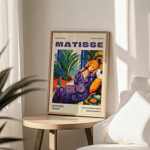 May include: Framed poster of Matisse's colorful artwork featuring a reclining woman in purple patterned clothing with orange head wrap, surrounded by potted plants. Displayed on light wood round side table next to beige upholstered chair. Warm natural lighting creates soft shadows on white walls. Exhibition-style poster shows "MATISSE" in purple letters with "06200 NICE, FRANCE" text below the vibrant fauve-style painting.