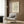 May include: Cream-colored poster with green coral or seaweed-like abstract shapes displayed in thin white frame on wood-paneled wall. Modern beige armchair with geometric patterned pillow, gold floor lamp with white shade, and dark round side table with open book visible in contemporary living room setting.