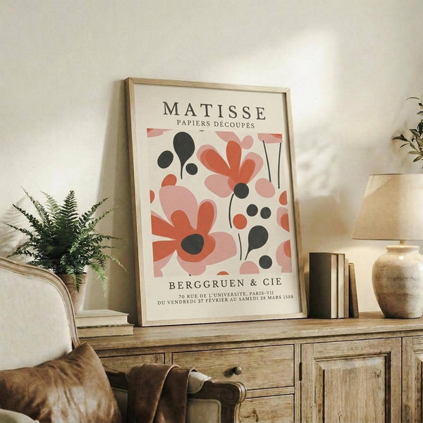May include: Framed Matisse exhibition poster with coral pink and black cut-out style botanical shapes displayed on rustic wooden sideboard. Scene shows neutral living room with cream armchair, fern plant, ceramic vase, stacked books, and warm natural lighting creating cozy home decor styling.