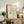 May include: Framed botanical poster with sage green coral-like cutout design on cream background, displayed on rustic wooden sideboard alongside fern plant, books, ceramic vase, and brown leather chair in neutral-toned living room setting
