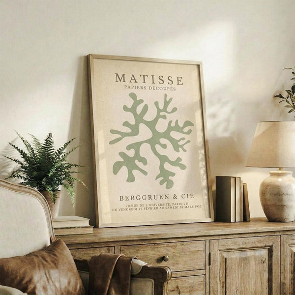 May include: Framed botanical poster with sage green coral-like cutout design on cream background, displayed on rustic wooden sideboard alongside fern plant, books, ceramic vase, and brown leather chair in neutral-toned living room setting