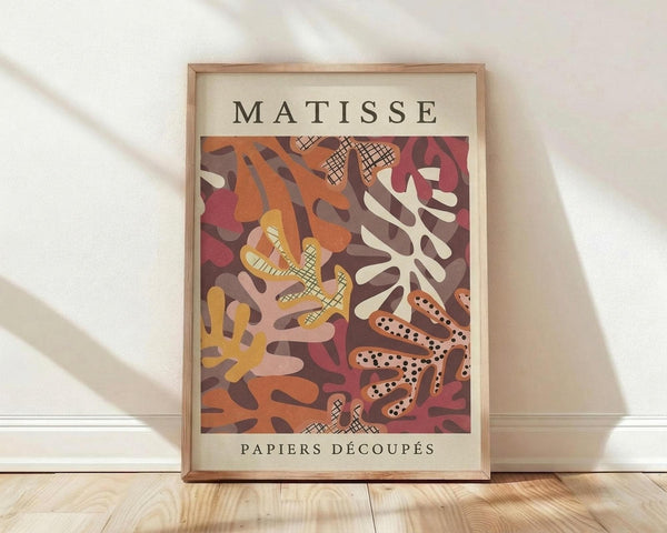 May include: Framed art print in light wood frame leaning against white wall on wooden floor. Poster features abstract cut-out shapes in warm tones of orange, burgundy, yellow and cream with crosshatch patterns and organic coral-like forms. Text reads "MATISSE" at top and "PAPIERS DÉCOUPÉS" at bottom on cream background. Soft natural lighting creates diagonal shadows across the scene.