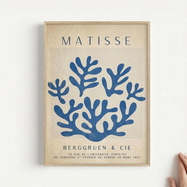 Framed poster print featuring a blue coral-like abstract shape on cream textured background. Text reads "MATISSE" at top and "BERGGRUEN & CIE" below the artwork, with French gallery exhibition details at bottom. Light wood frame against white wall with partial hand visible in corner for scale reference.